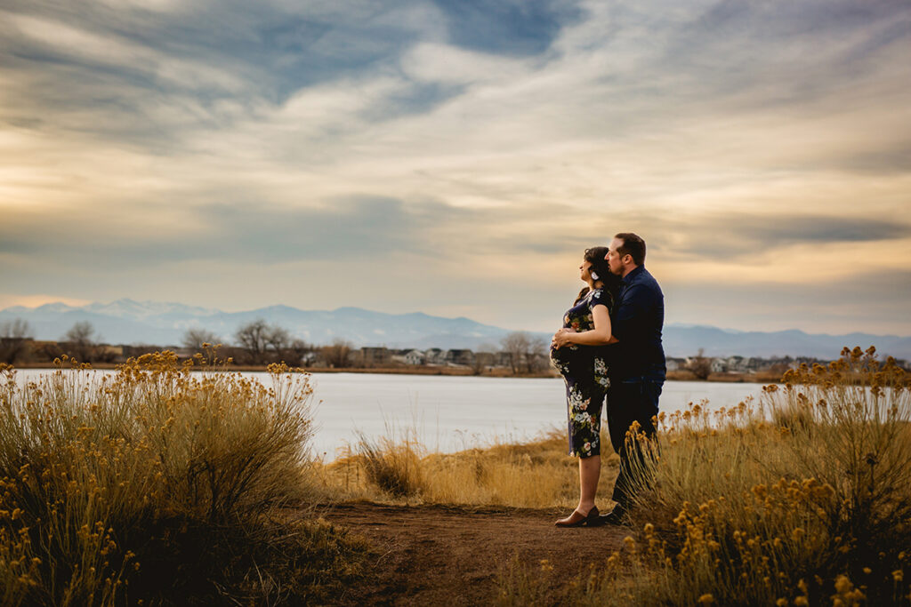A couple looks at the beautiful sunset over the mountains as they pose for a maternity photo near a lake in Loveland, Colorado